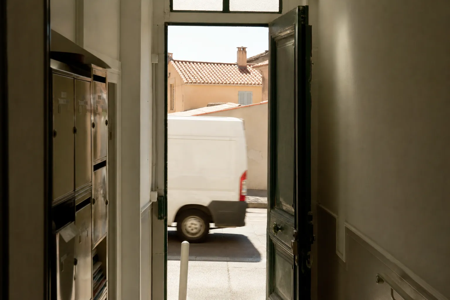 Porte d’entrée d’une maison marseillaise vue depuis le couloir, entrebâillée sur la rue claire, camionnette blanche floue au bord du trottoir