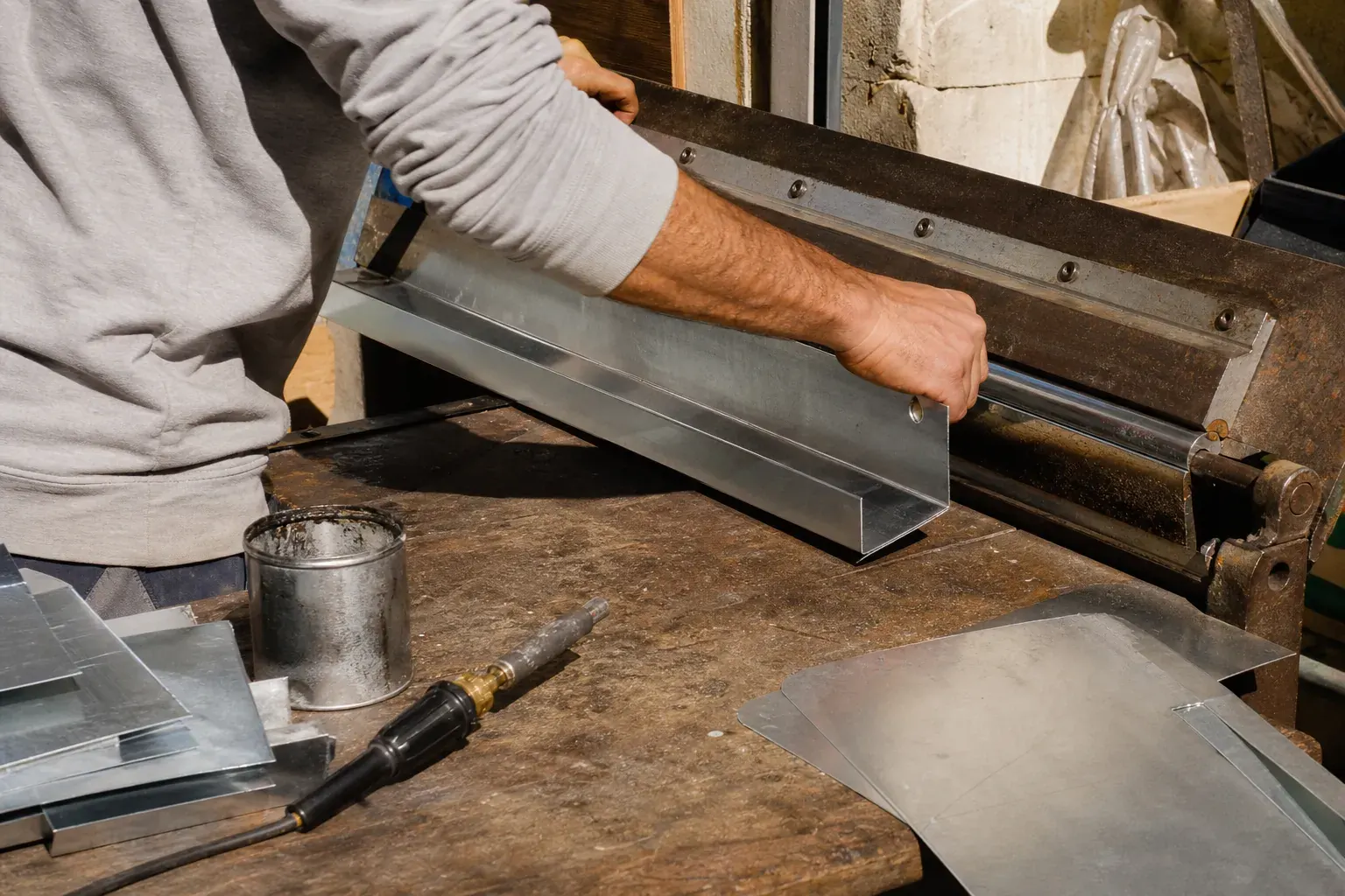 Mains de zingueur pliant une feuille de zinc sur une plieuse pour former un profil de gouttière, fer à souder et étain posés à côté