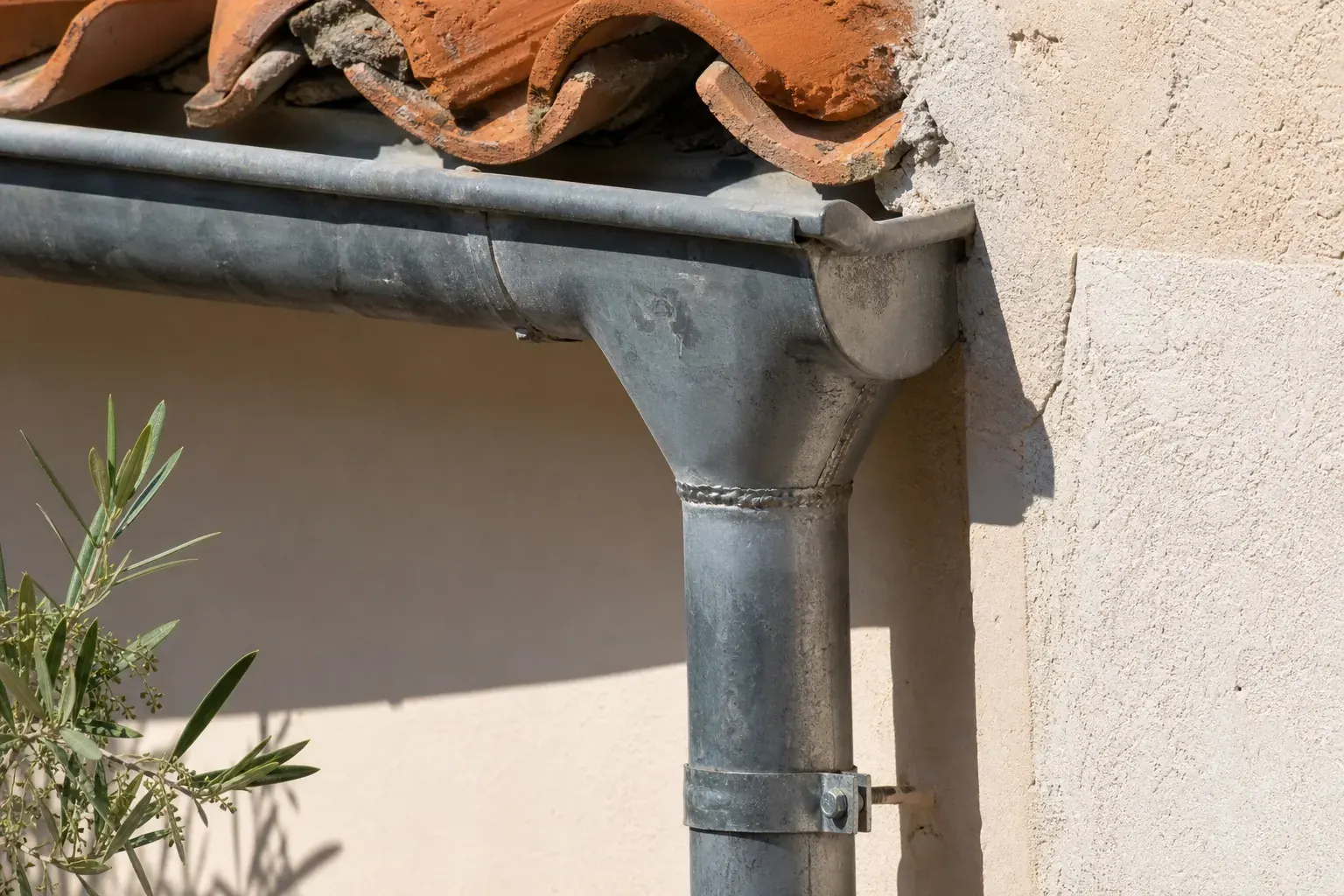 Gros plan d’une gouttière de zinc et de sa naissance soudée alimentant une descente ronde fixée sur un mur crème