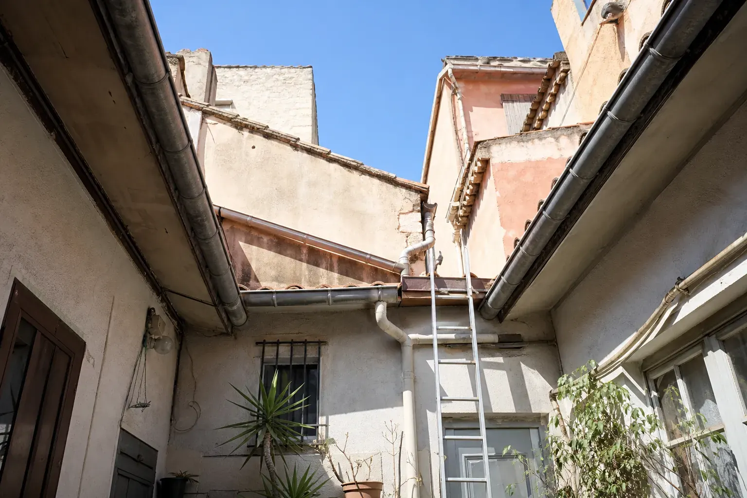 Cour intérieure d’un immeuble ancien vue d’en bas : rives de toit sur les quatre côtés, gouttière de zinc en périphérie, échelle appuyée