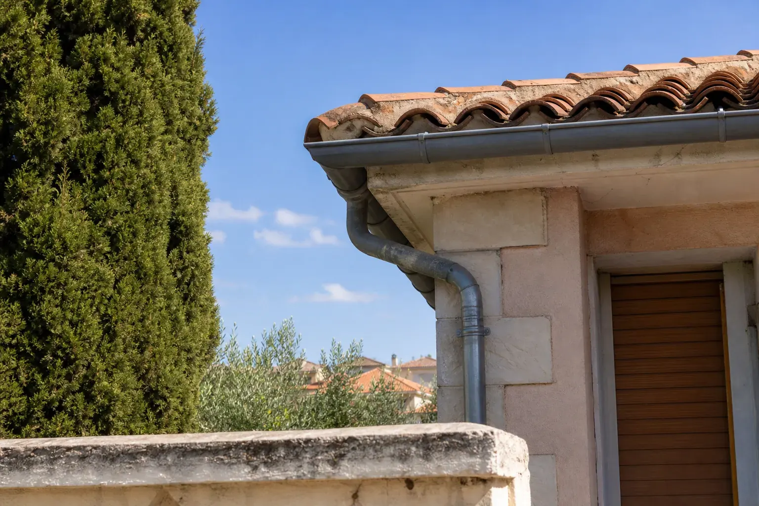 Égout de toit d’un pavillon avec sa gouttière pendante et sa descente, tuiles mécaniques au-dessus, cyprès au premier plan